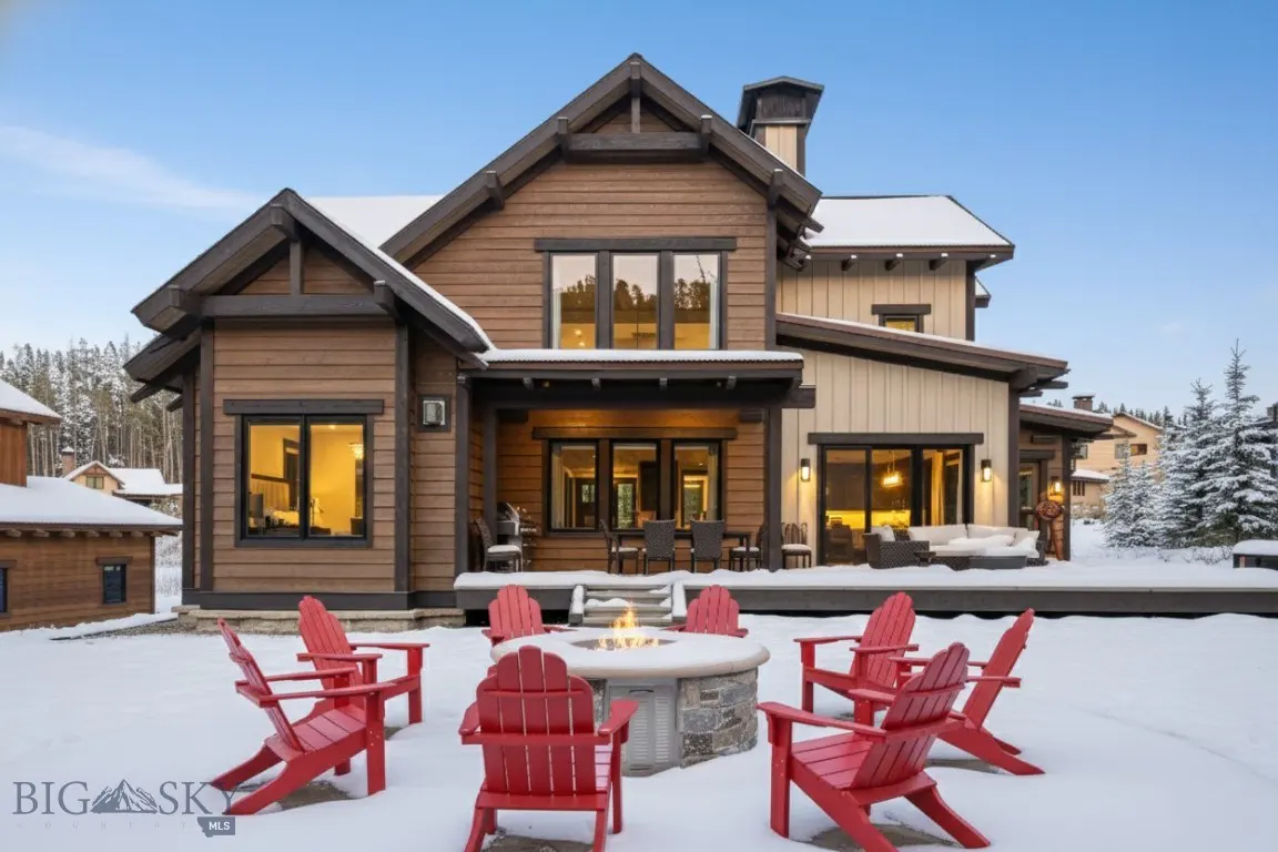 Exterior view of a two-story brown and beige house with a stone fire pit surrounded by red Adirondack chairs in the snow.