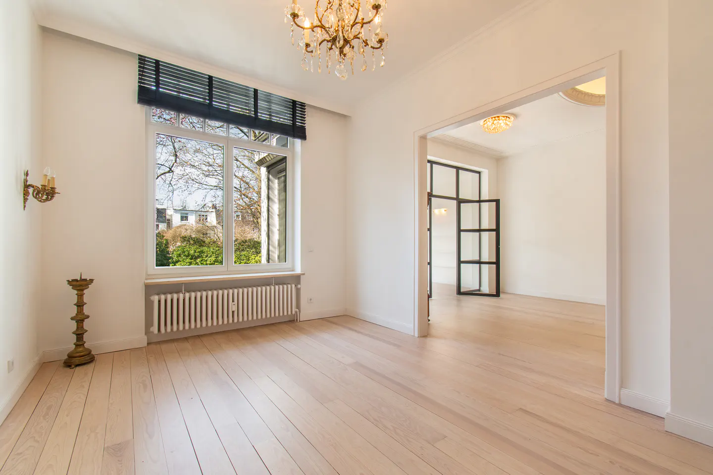 Bright, empty room with light wood floors, white walls, and a chandelier. A large window overlooks a green yard. An open doorway leads to another room.