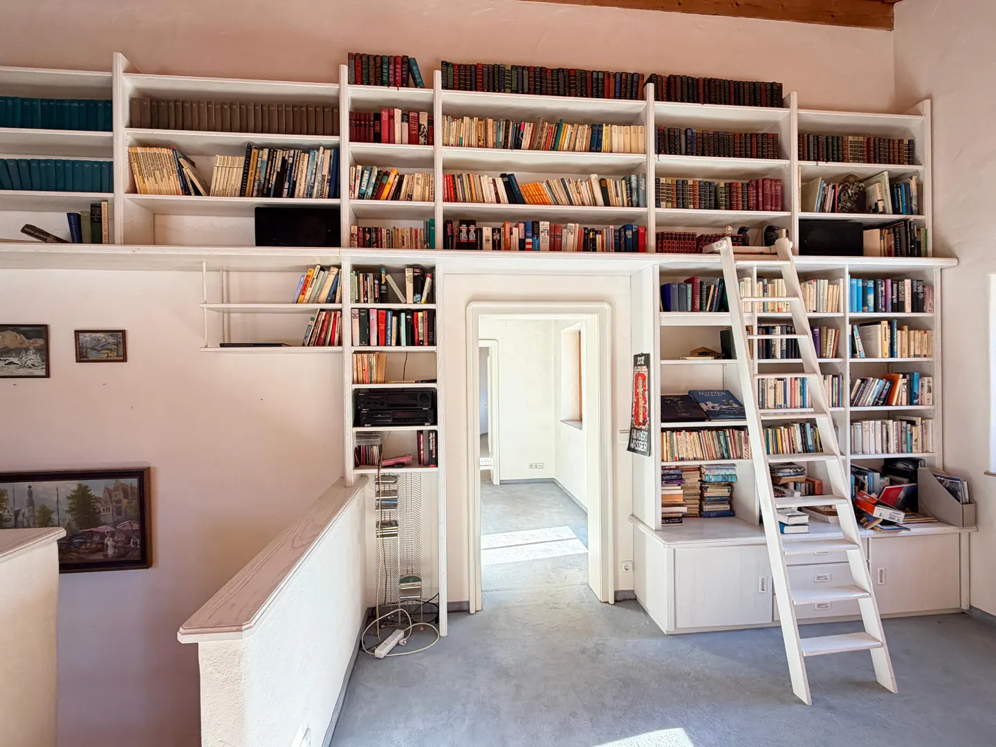 A white, floor-to-ceiling bookshelf filled with books, with a white ladder leaning against it. A doorway leads to another room.