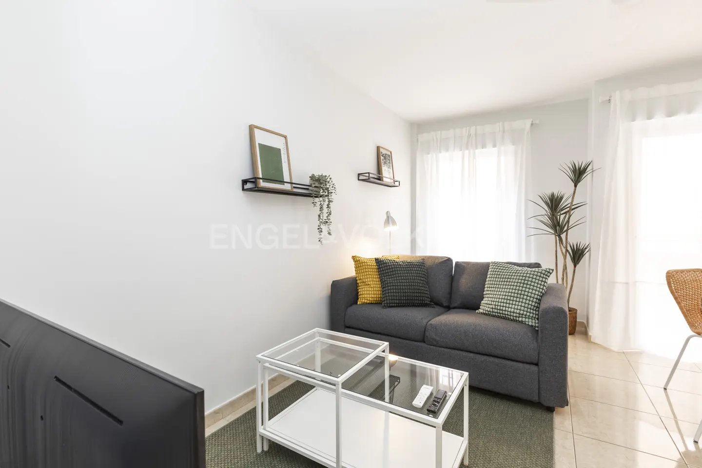 Living room with gray sofa, pillows, glass table, and white walls with shelves.