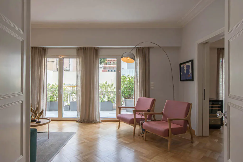Living room with herringbone wood floors, two pink chairs, and a curved floor lamp. Balcony with plants visible through glass doors.