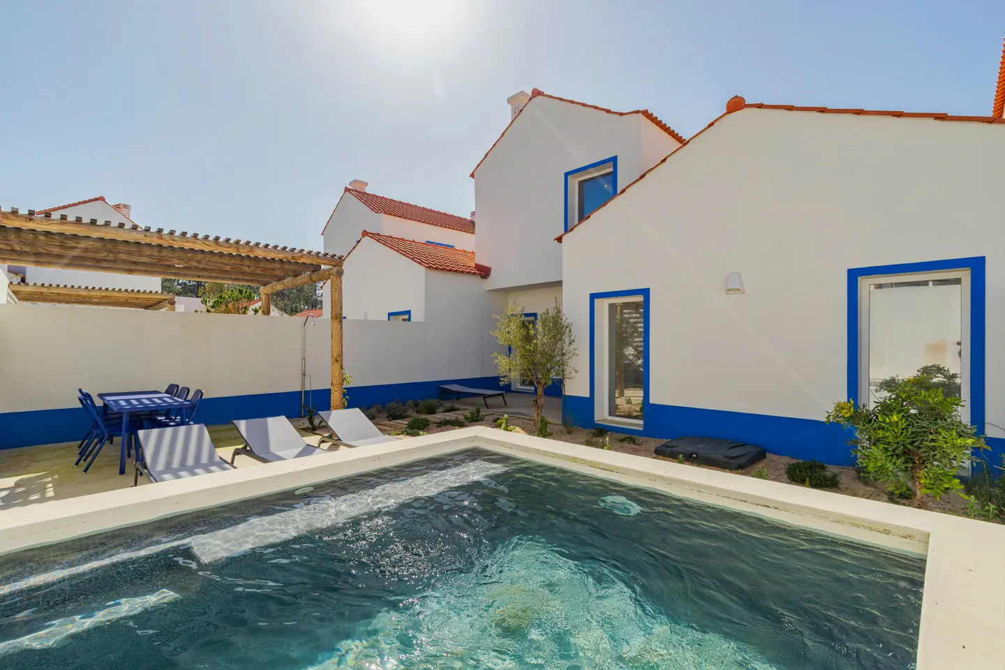 Exterior view of a white house with a red roof and blue trim, a pool, and outdoor furniture.