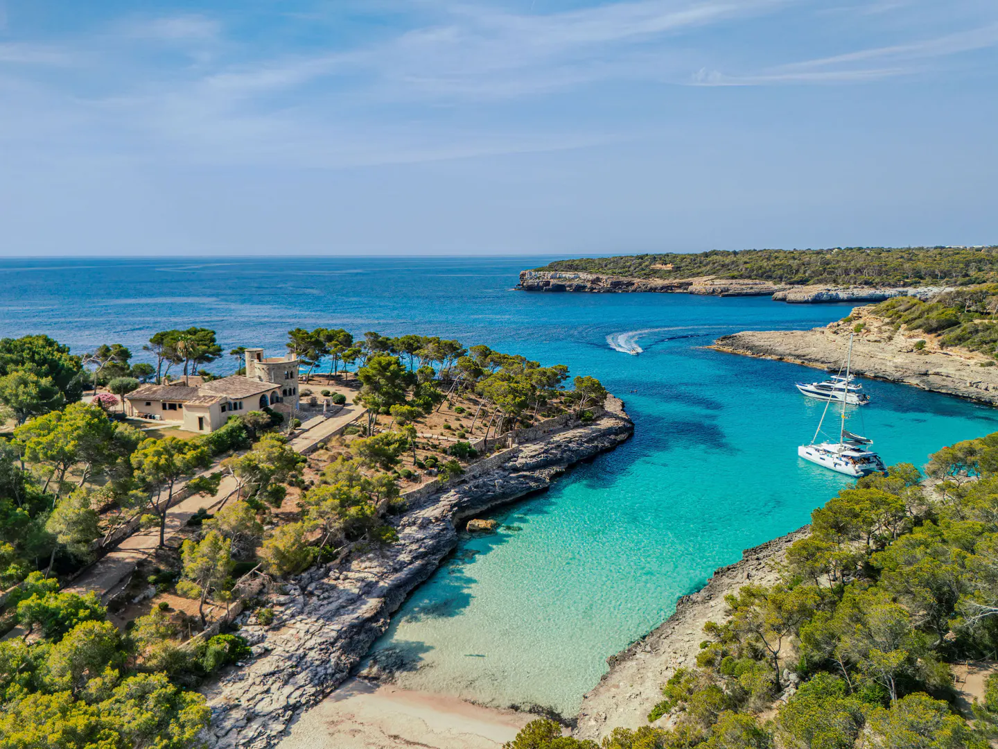 Aerial view of a turquoise bay with boats, a sandy beach, and a villa on a tree-lined coast under a blue sky.
