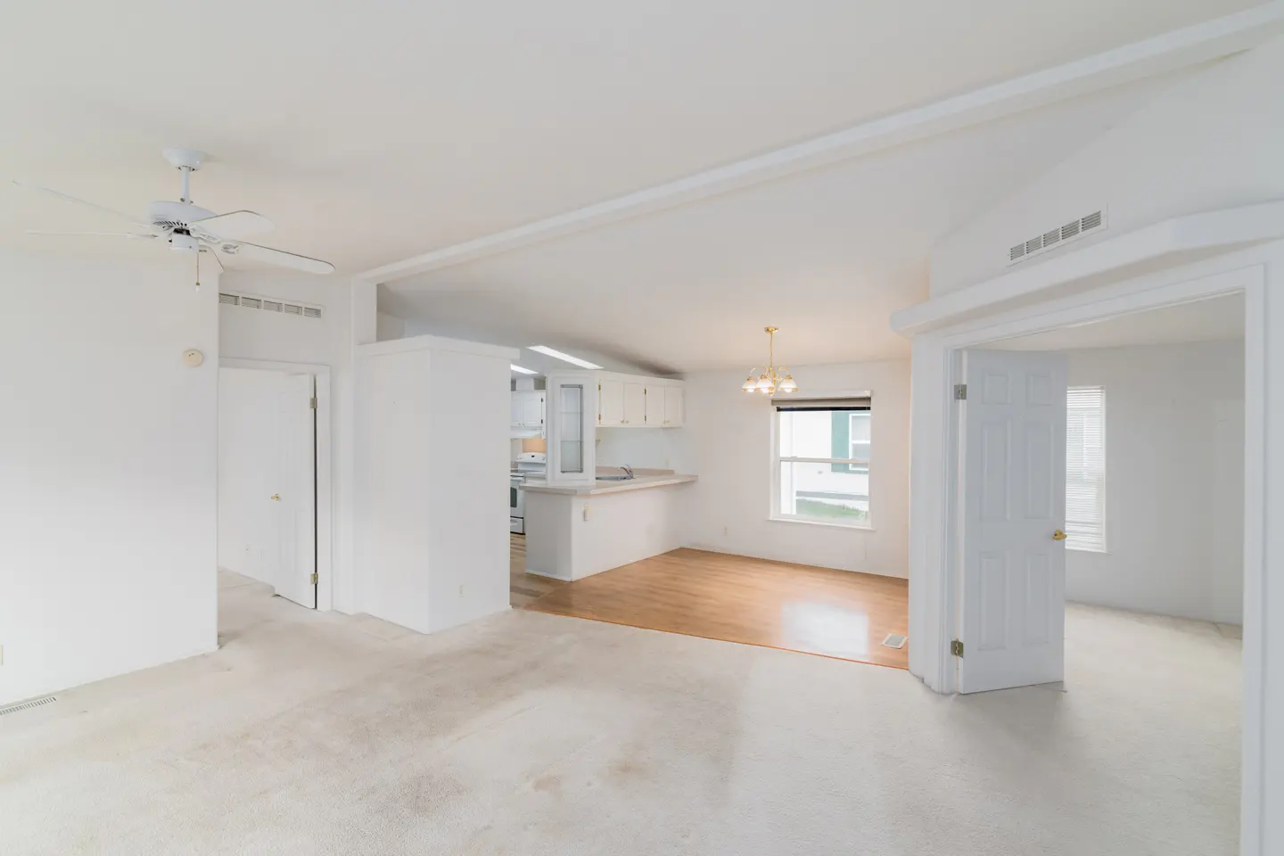 Bright, open-concept living space with white walls, carpet, and wood flooring. A kitchen with white cabinets is visible in the background.