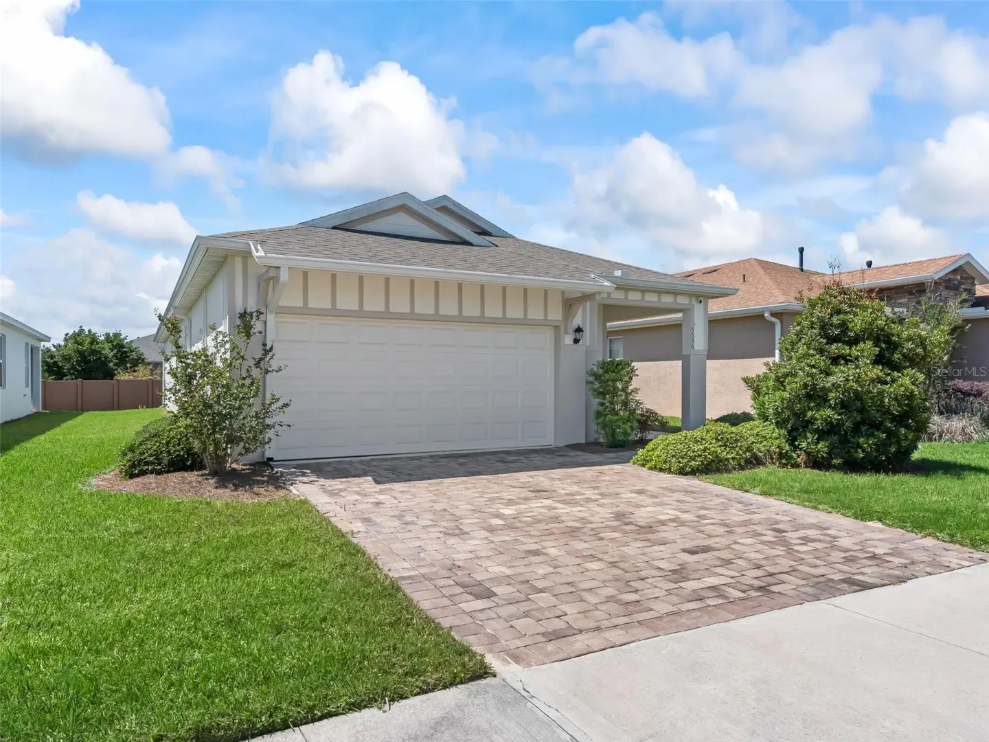 Single-story home with a white garage door, tan siding, and a brick driveway. Green lawn and blue sky with clouds.