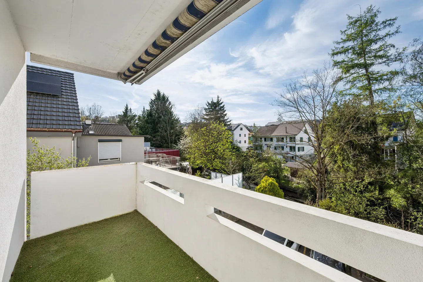 Balcony view with artificial grass, white walls with rectangular openings, and a striped awning. Trees and houses are visible in the background.