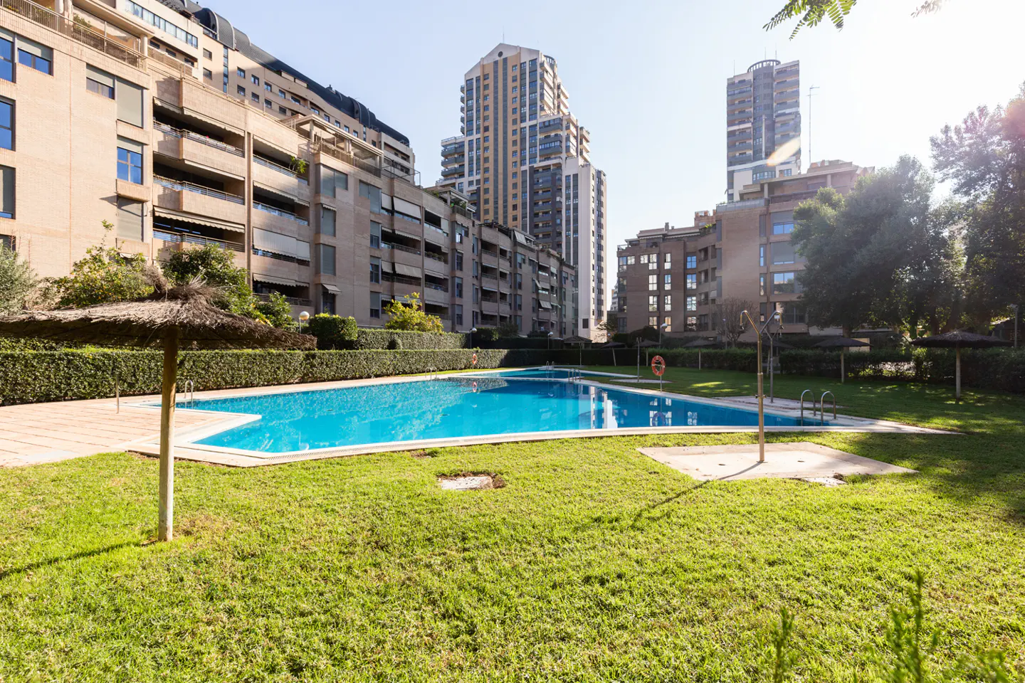 Outdoor pool with blue water, surrounded by green grass and buildings in the background. A straw umbrella is on the left.