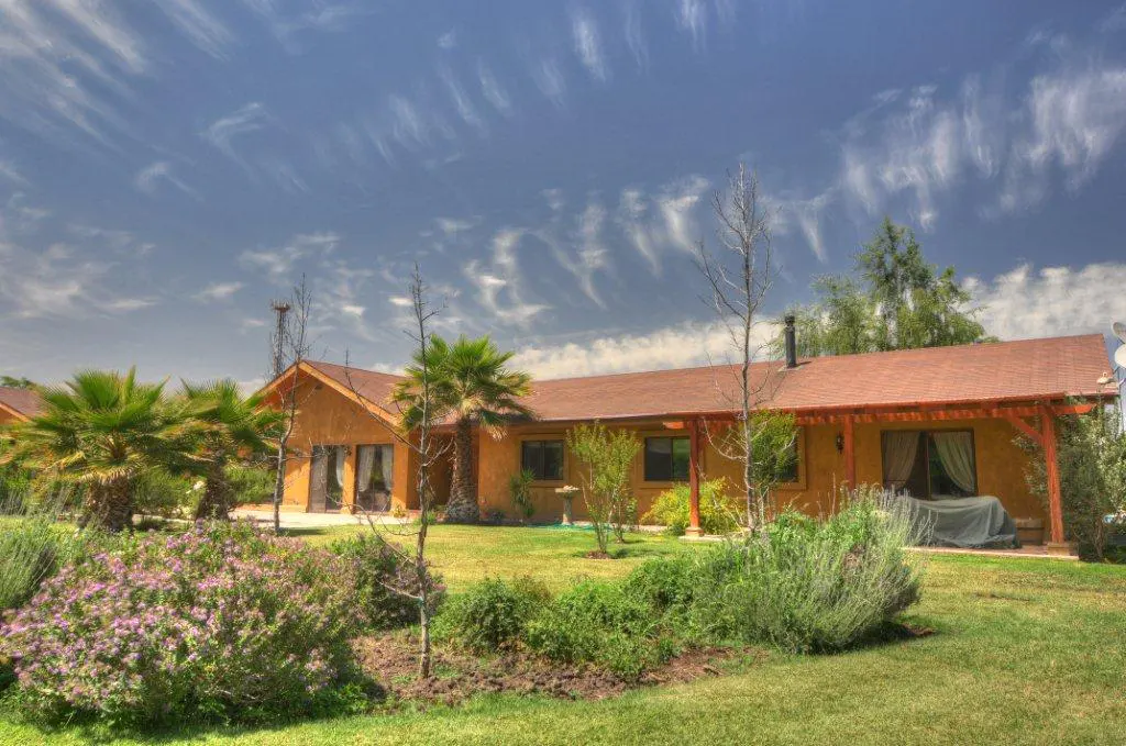 Exterior view of a tan house with a brown roof, green lawn, and palm trees under a blue sky with wispy clouds.