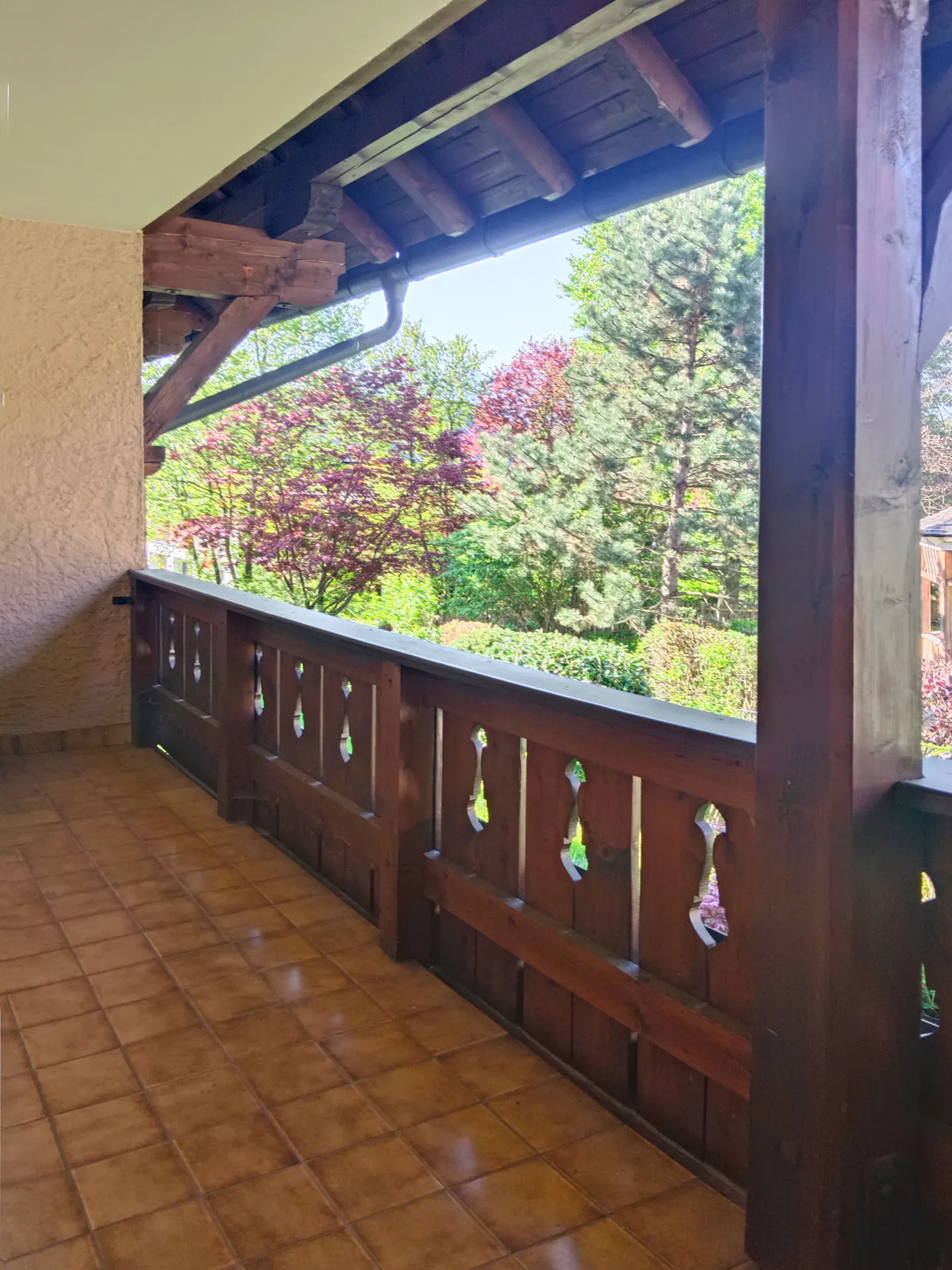 Covered porch with brown tile floor and carved wood railing overlooking a lush green landscape with trees.