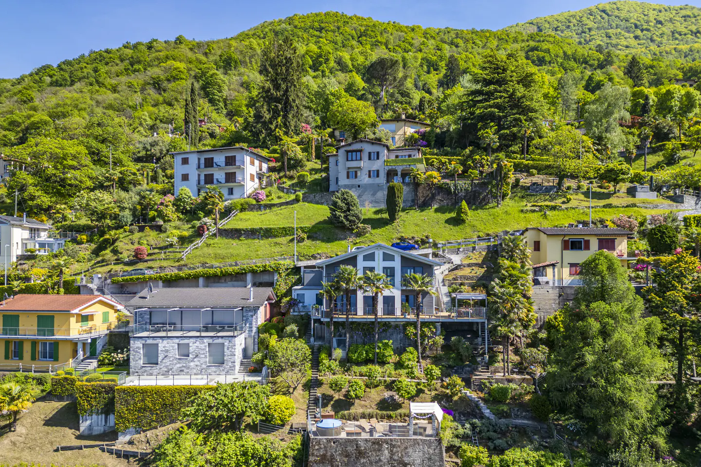 Multi-level hillside homes with green lawns and trees under a blue sky.