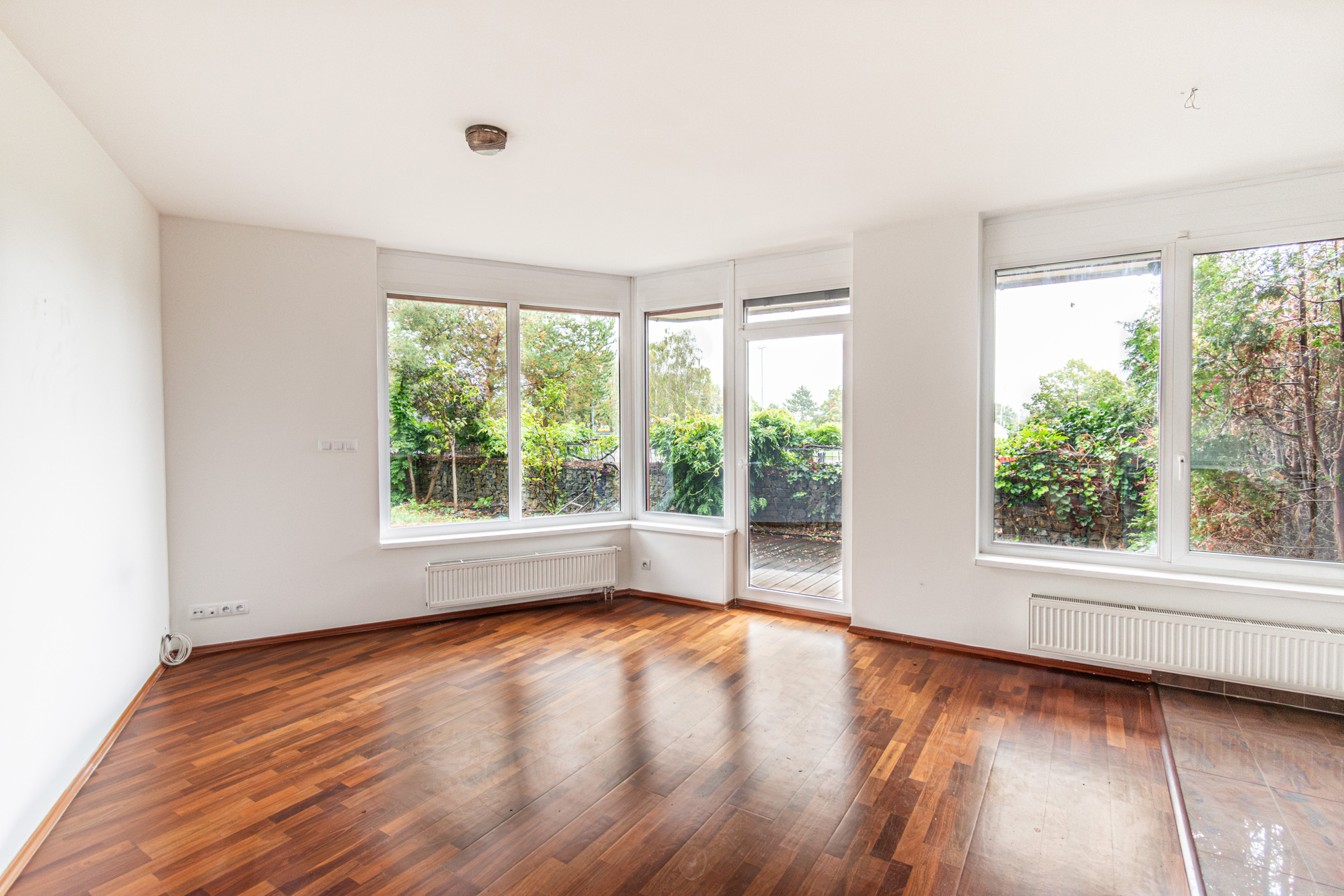 Bright, empty room with dark wood floors, white walls, large windows, and a glass door leading to a patio. [AI-generated text]