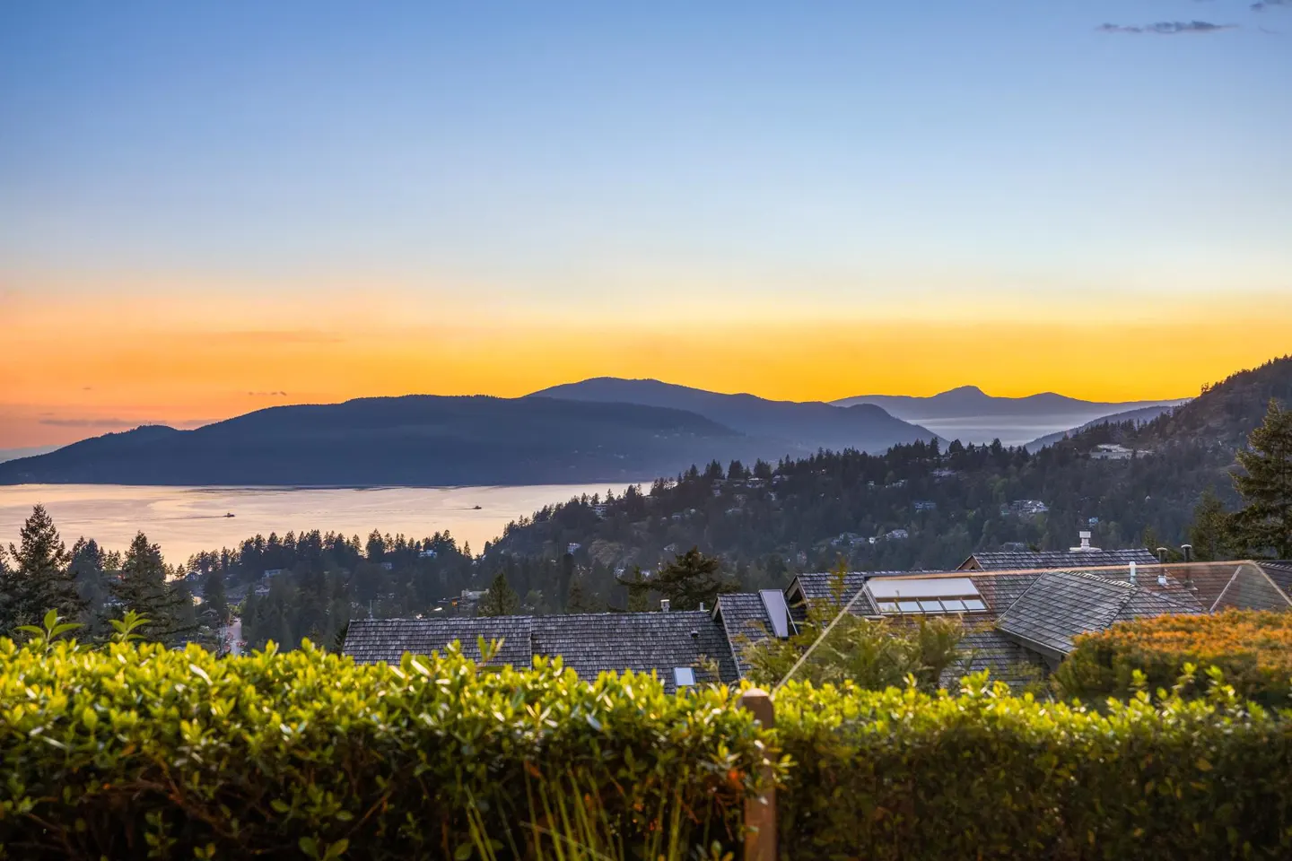Sunset view of mountains and ocean, with rooftops and green hedges in the foreground. The sky is orange and blue.