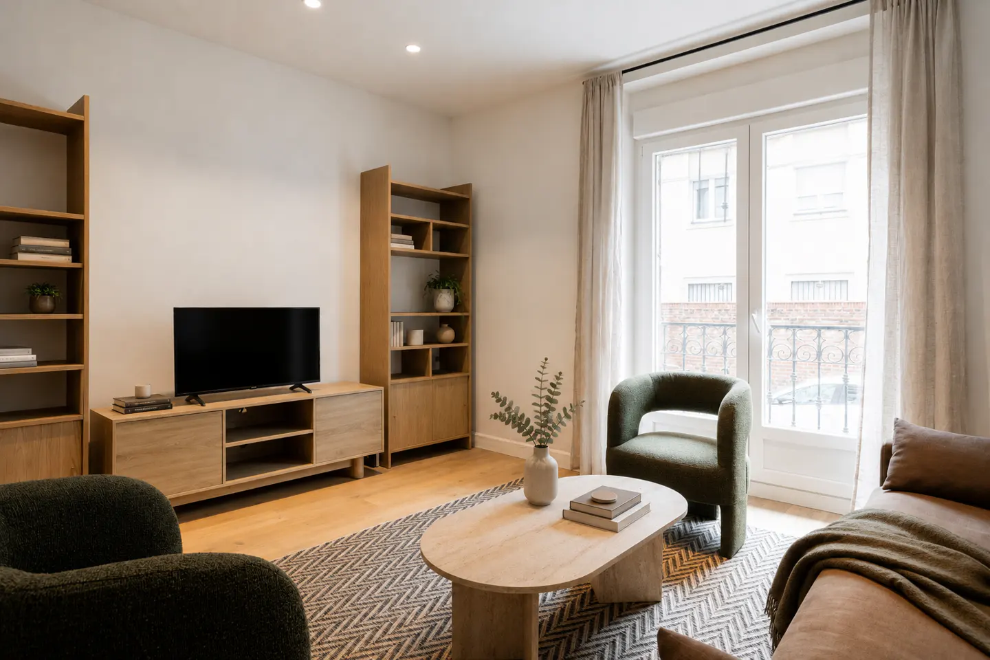 Living room with light wood floors, bookshelves, TV, and green chairs. A patterned rug sits under a light wood coffee table. Curtains frame a window.