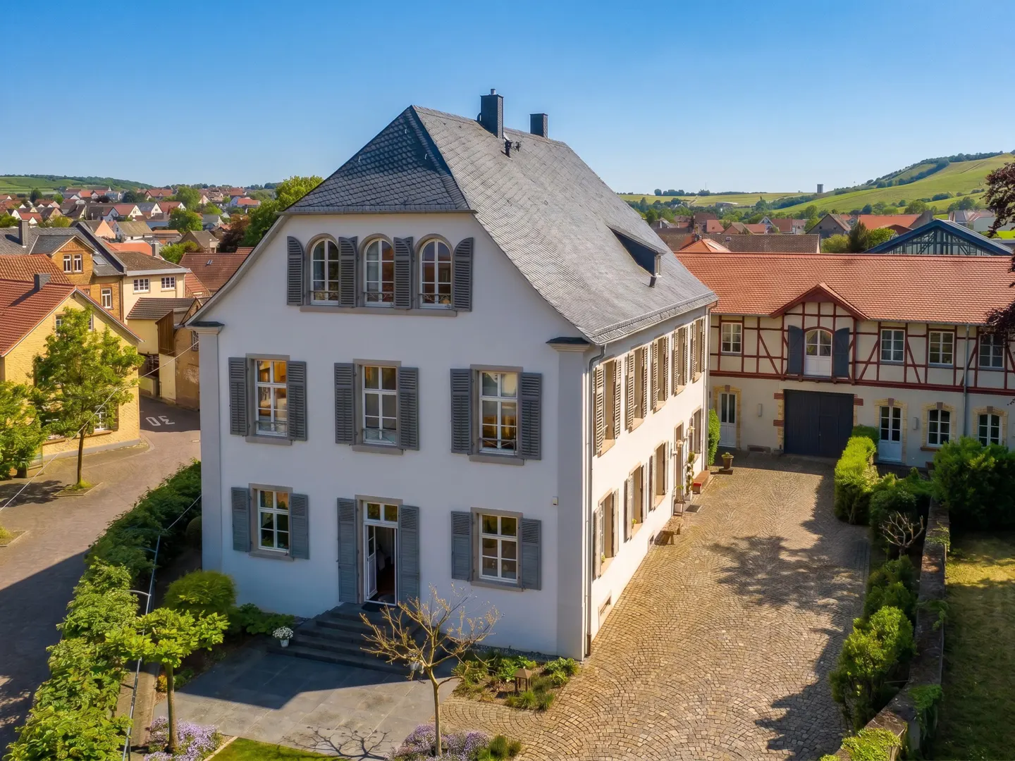 Three-story white house with gray shutters and roof, cobblestone driveway, and green landscaping.