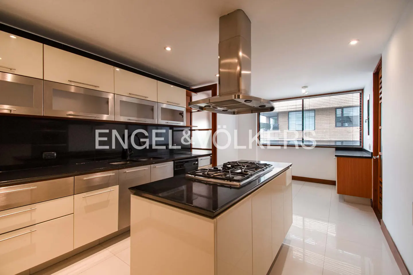 A modern kitchen with cream cabinets, black countertops, and a stainless steel island range hood. The floor is white tile.