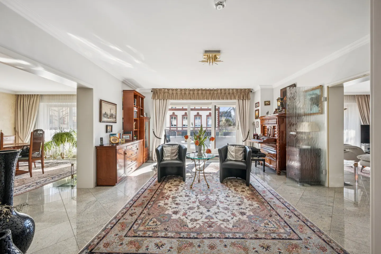 Bright living room with marble floors, a large rug, two black armchairs, and a glass table with flowers. A piano and wooden furniture are visible.