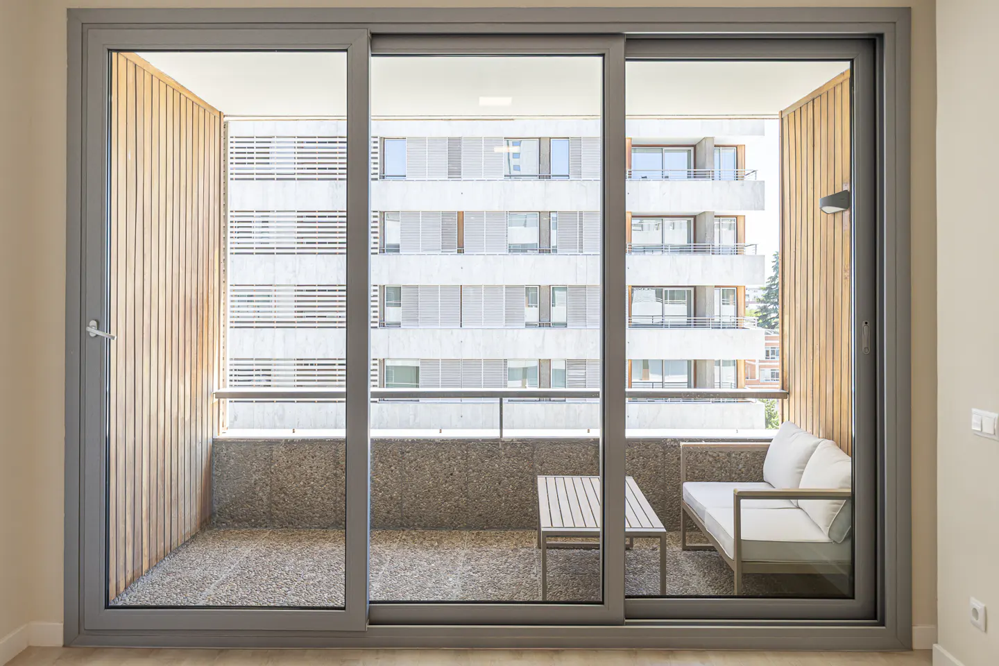 Large sliding glass doors open to a balcony with a small table, a white sofa, and wood-paneled walls overlooking a city.