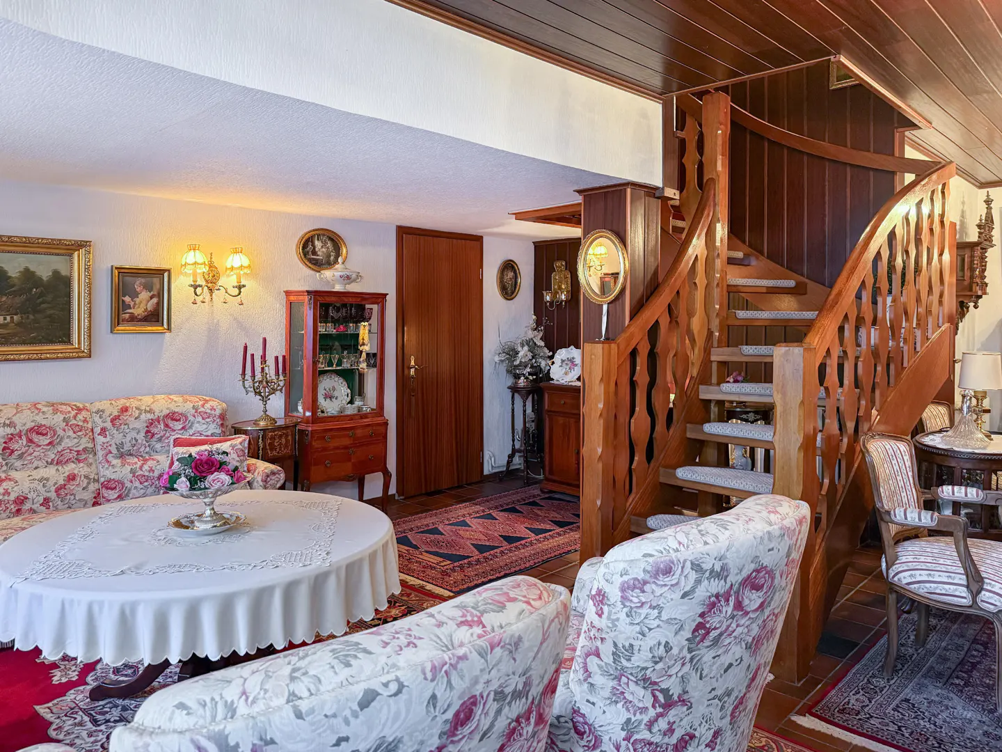 Cozy living room with floral sofas, round table, wood staircase, and antique decor. Warm lighting and wood accents create a traditional ambiance.