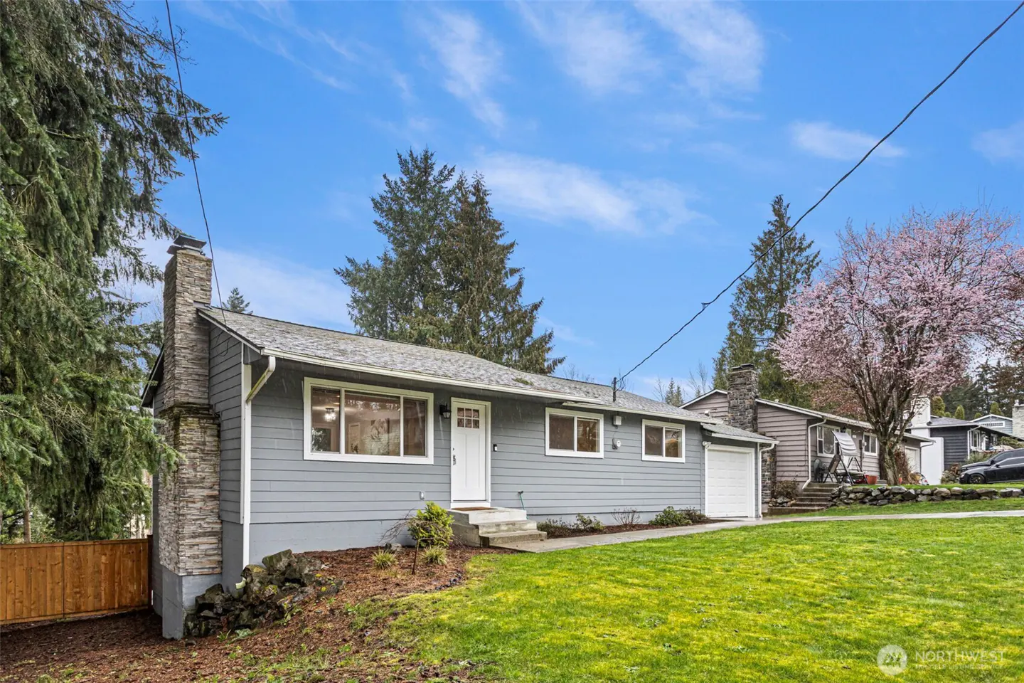 Single-story gray house with a stone chimney, white trim, and a front lawn under a blue sky with a blooming cherry tree.