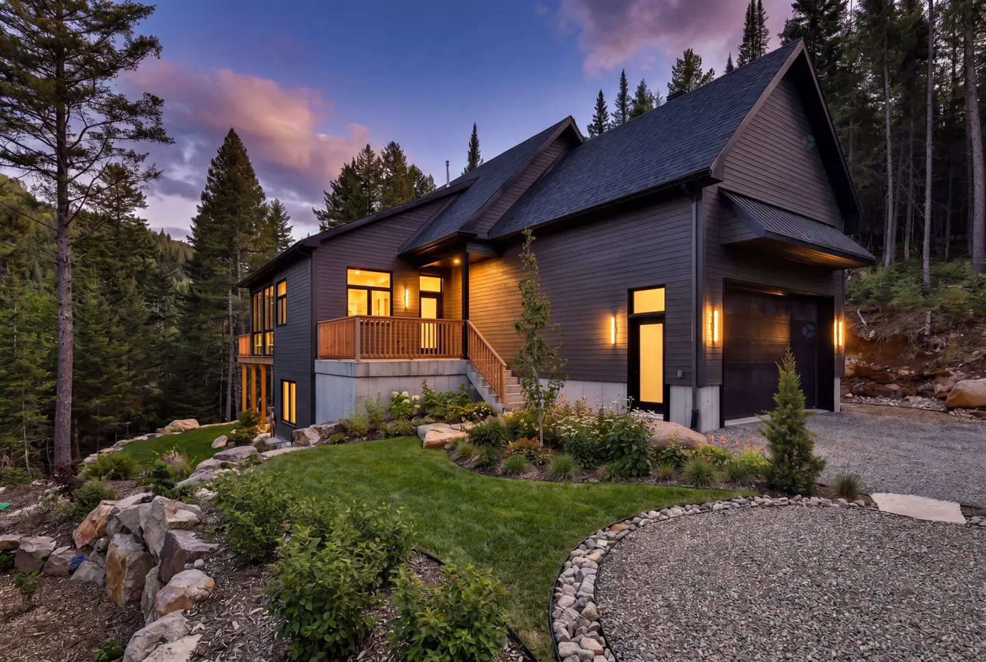 Exterior view of a modern, two-story house with a dark gray facade, a wooden deck, and a garage, surrounded by lush greenery and tall trees.