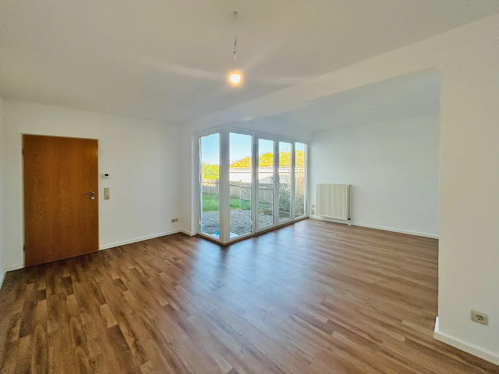 Bright, empty room with wood floors, white walls, and a glass door to a backyard. A wooden door is on the left.
