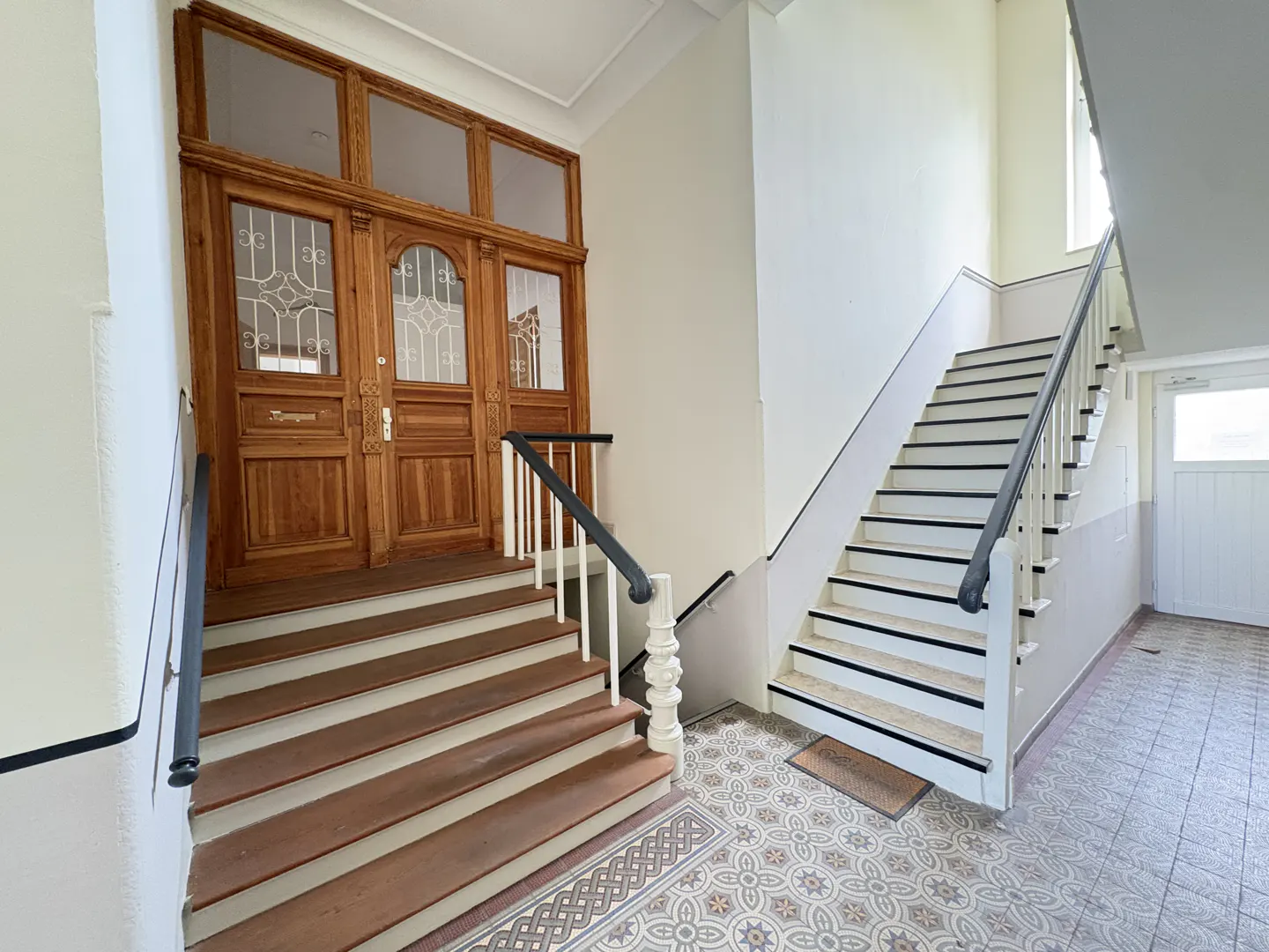 Interior view of a building entrance with wooden doors, stairs, and patterned tile flooring.