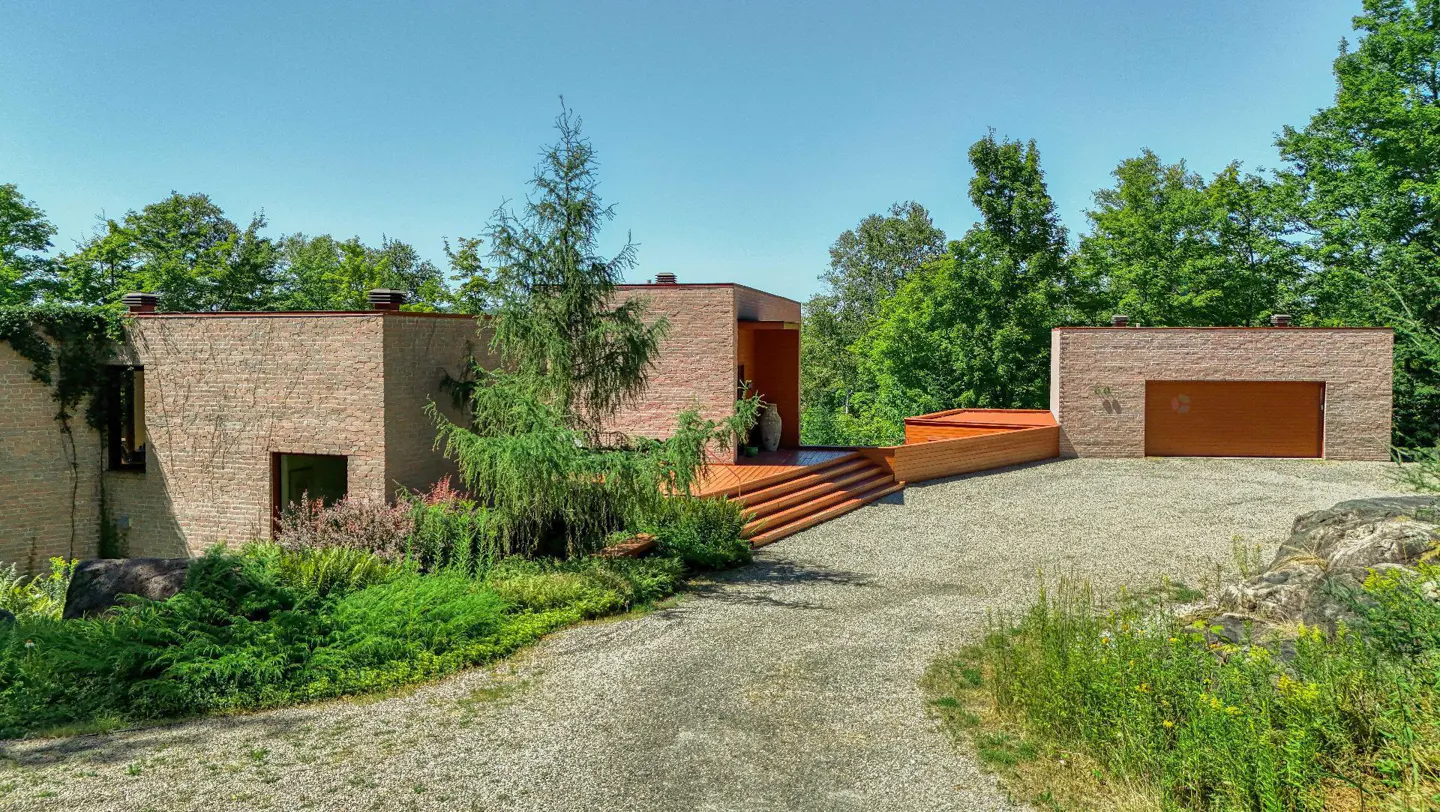 Exterior view of a modern, tan brick home with a wooden deck and garage, surrounded by lush greenery and a gravel driveway.
