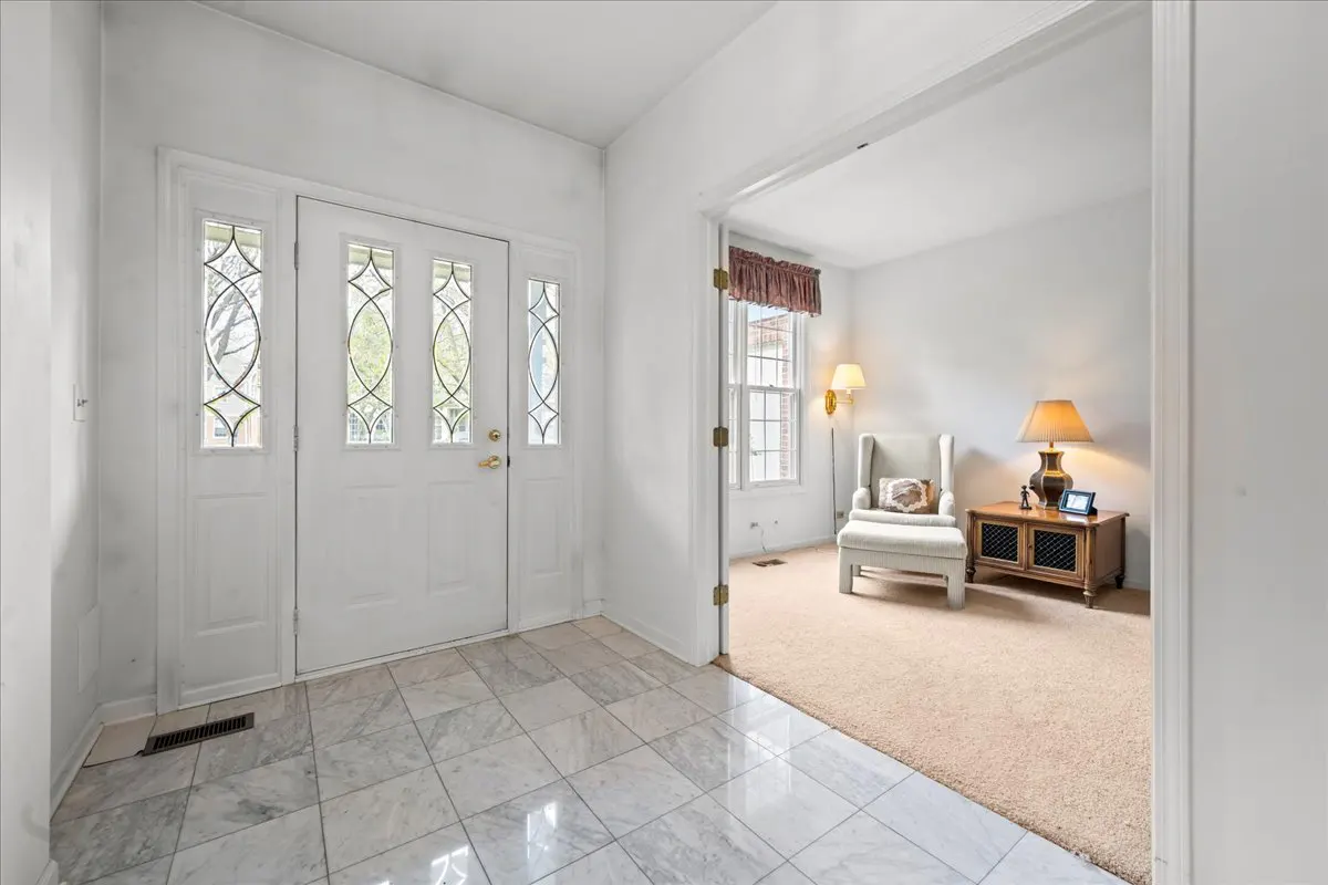 Foyer with white door and marble tile floor, leading to a living room with armchair, side table, and lamps.