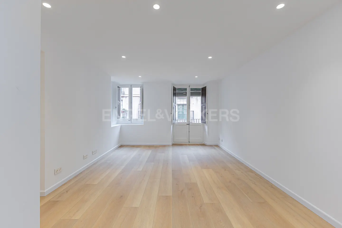 Empty white room with light wood floors, two windows with dark shutters, and a double door leading to a balcony.