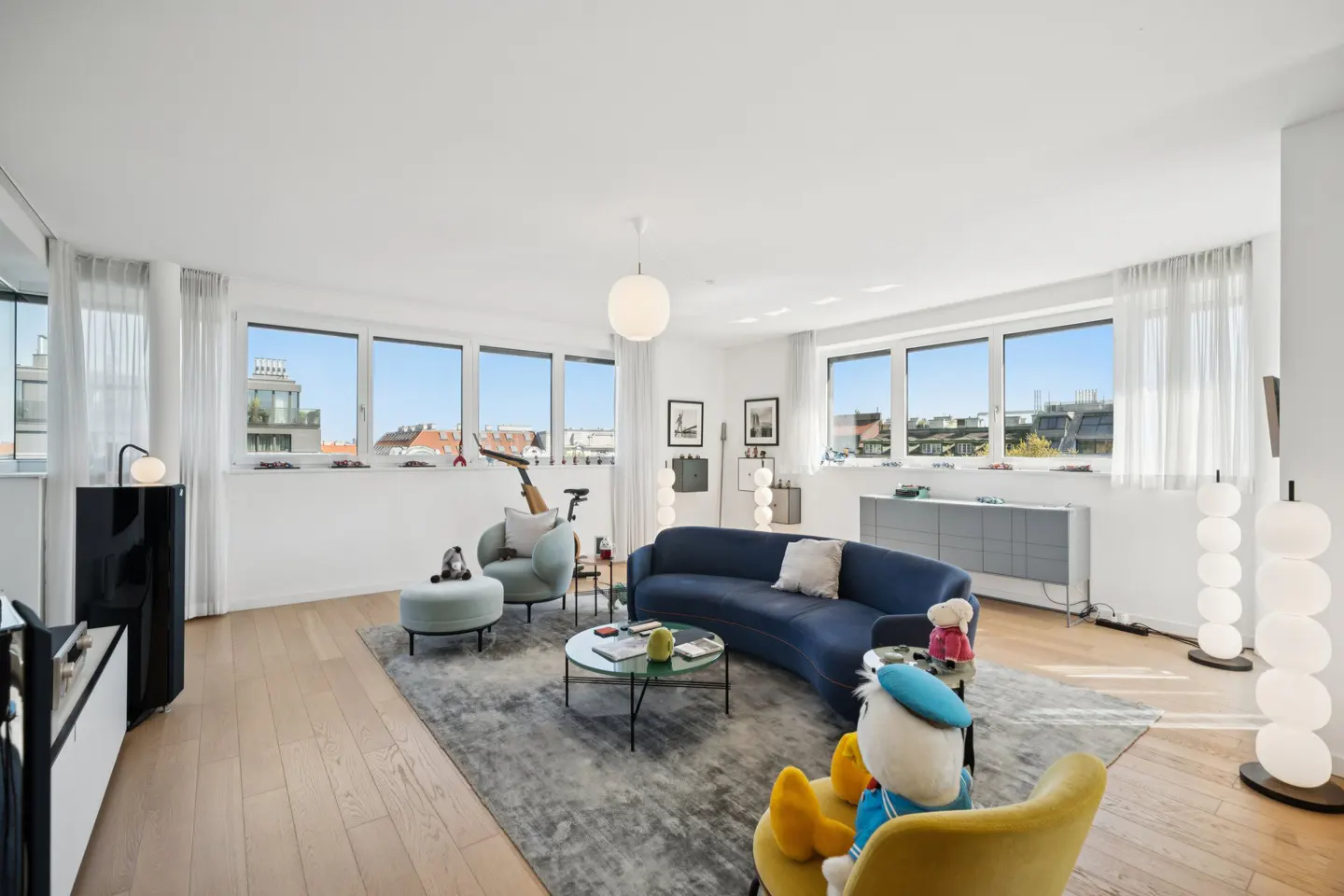 Bright, modern living room with wood floors, blue sofa, and large windows overlooking a city. A yellow chair with a Donald Duck plush is in the foreground.