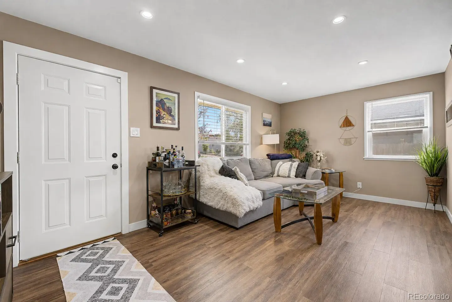 Living room with wood floors, a gray sectional sofa, and a bar cart with liquor bottles. A white door and patterned rug are on the left.