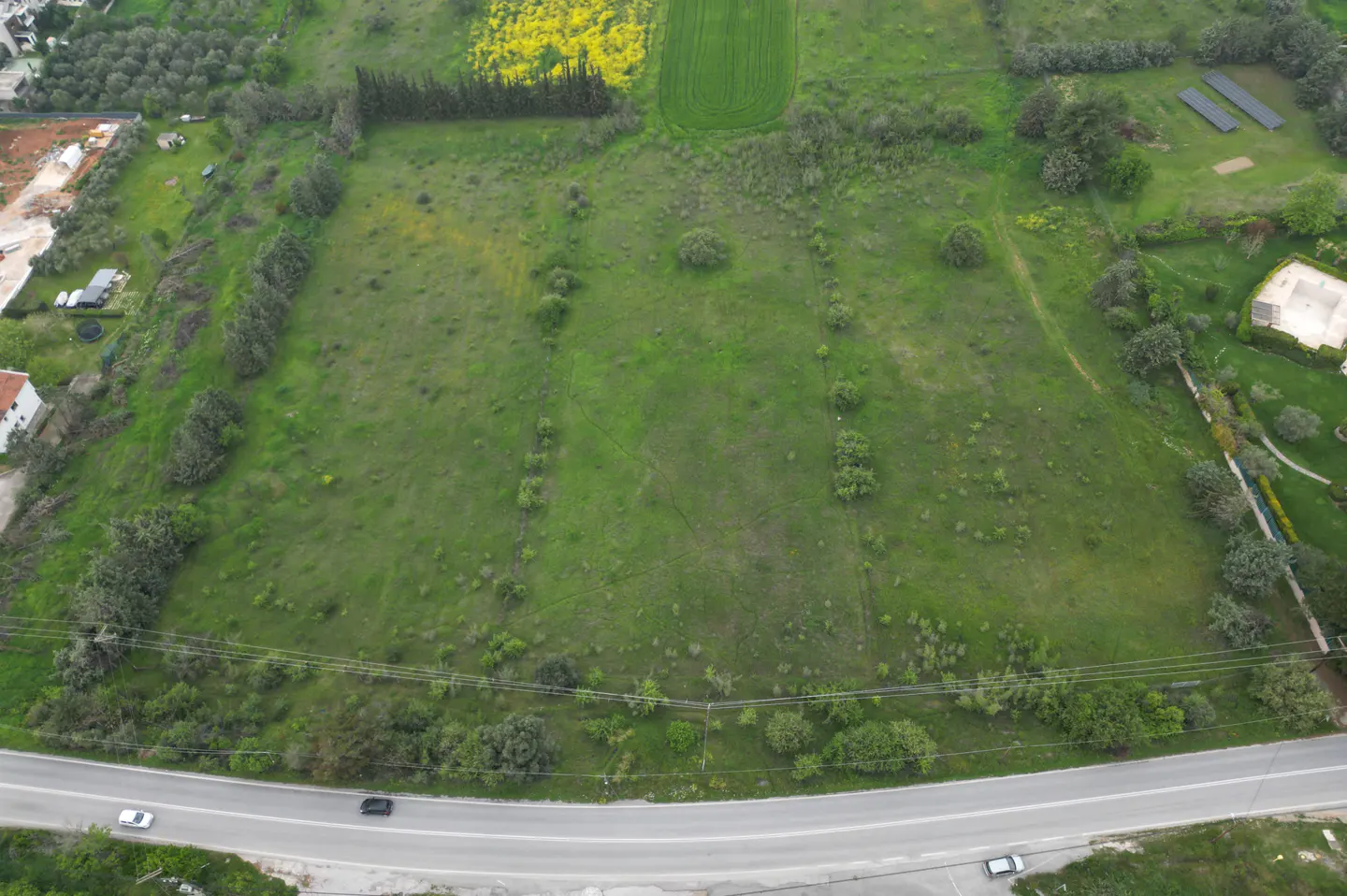 Aerial view of a large, green, grassy lot with trees, a road, and some buildings in the background.