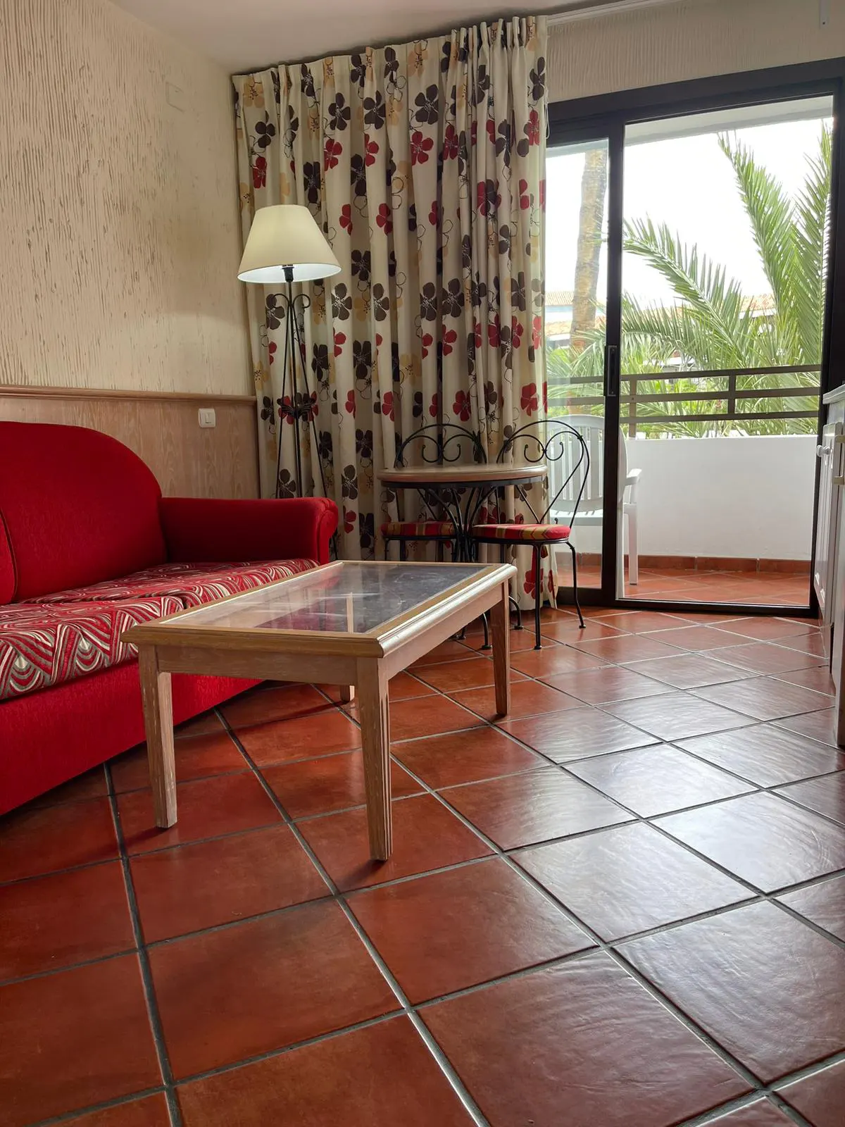 Living room with red sofa, wood table, and balcony view. Floral curtains frame the sliding glass door.