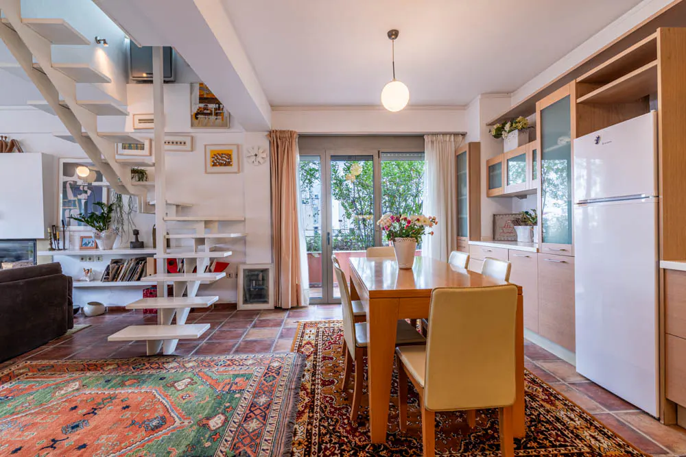 Dining area with wooden table, chairs, and white refrigerator, featuring a staircase and colorful rugs on a tiled floor.
