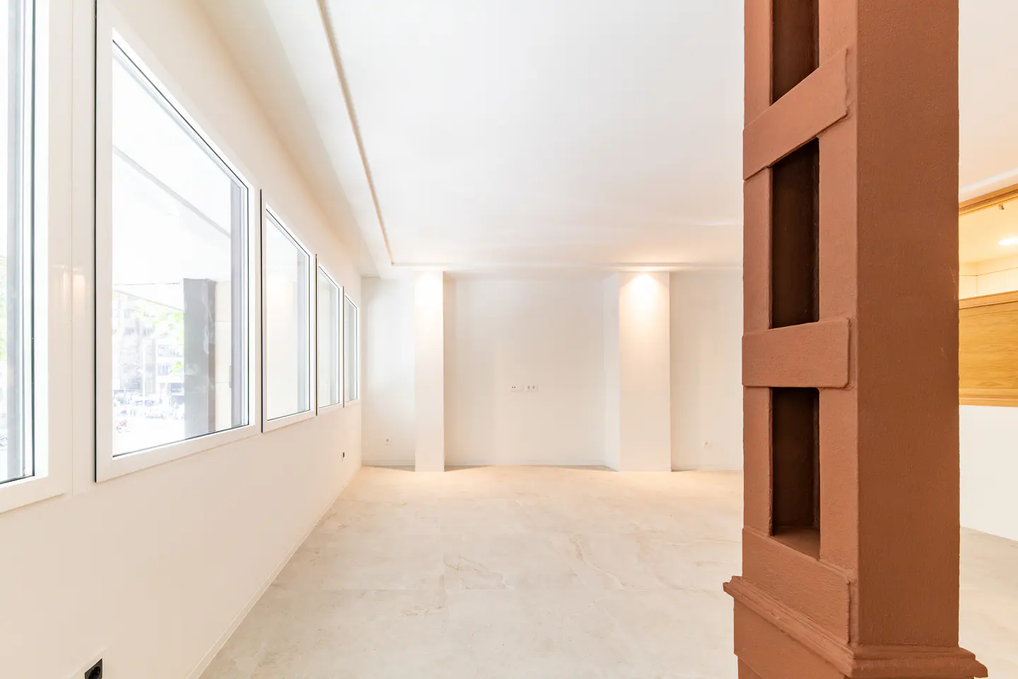 Bright, empty room with white walls, light tile floors, large windows on the left, and a decorative brown pillar.