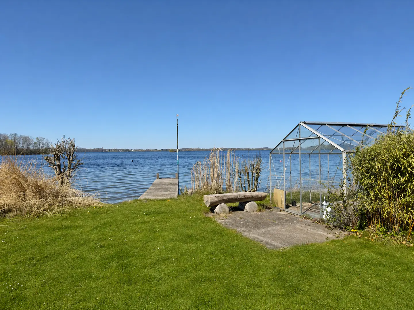 Lakeside property with a wooden dock, bench, and greenhouse on a sunny day. Blue water and sky.