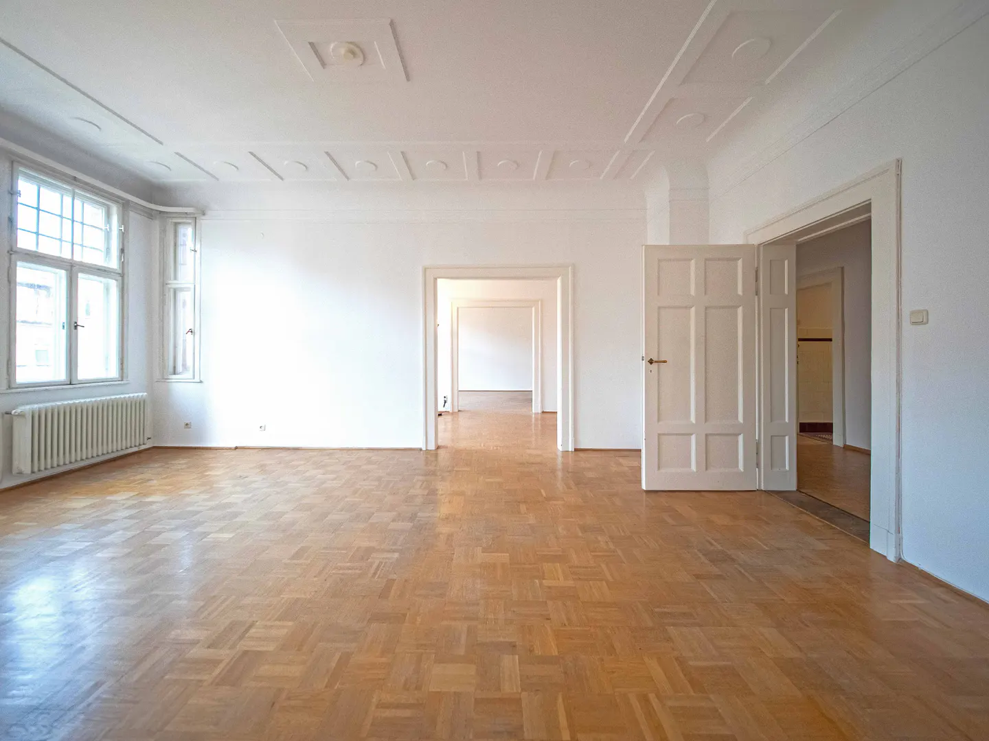 Bright, empty room with parquet floors, white walls, and decorative ceiling. Windows on the left, open doors lead to other rooms.