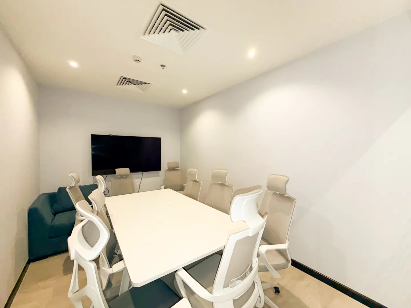 Small meeting room with a white table, eight ergonomic chairs, a wall-mounted TV, and a teal sofa against a white wall.