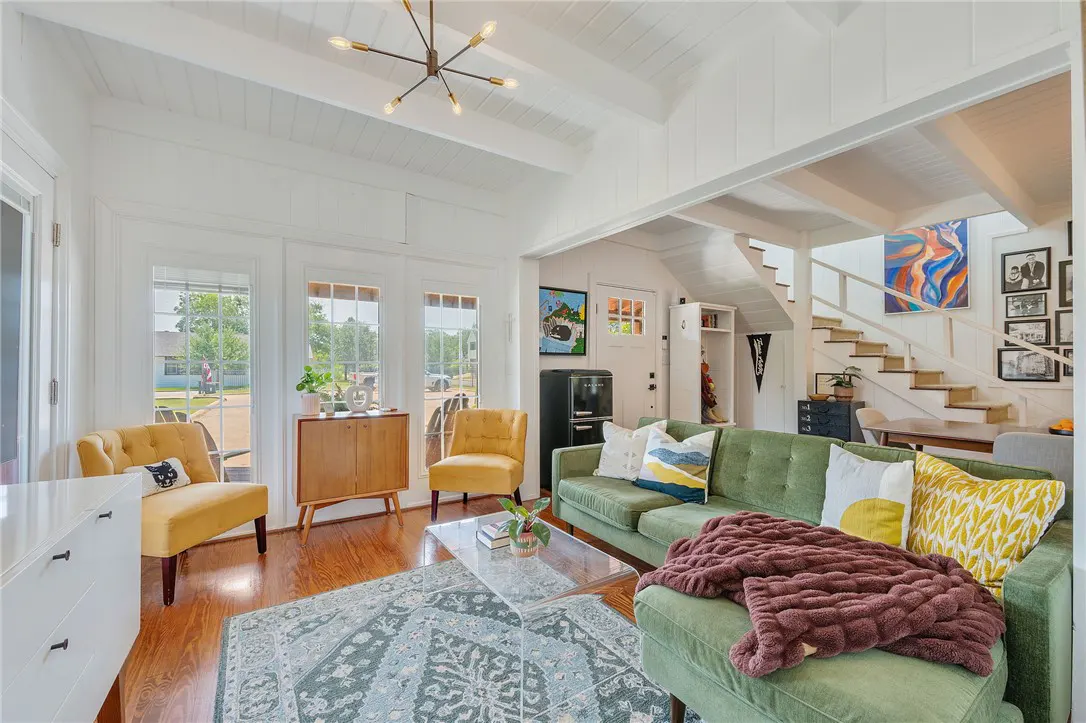 Bright living room with hardwood floors, a green sofa, yellow chairs, and a patterned rug. Stairs lead to the second floor.