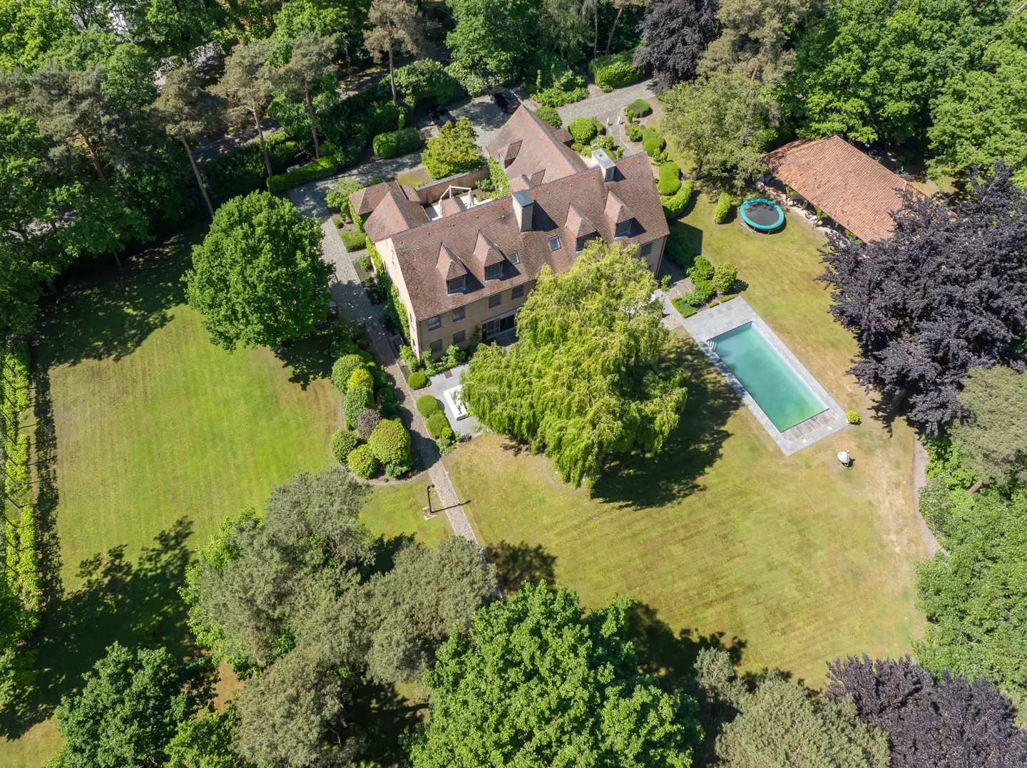 Aerial view of a large tan house with a brown roof, a pool, and a trampoline surrounded by green trees and grass.