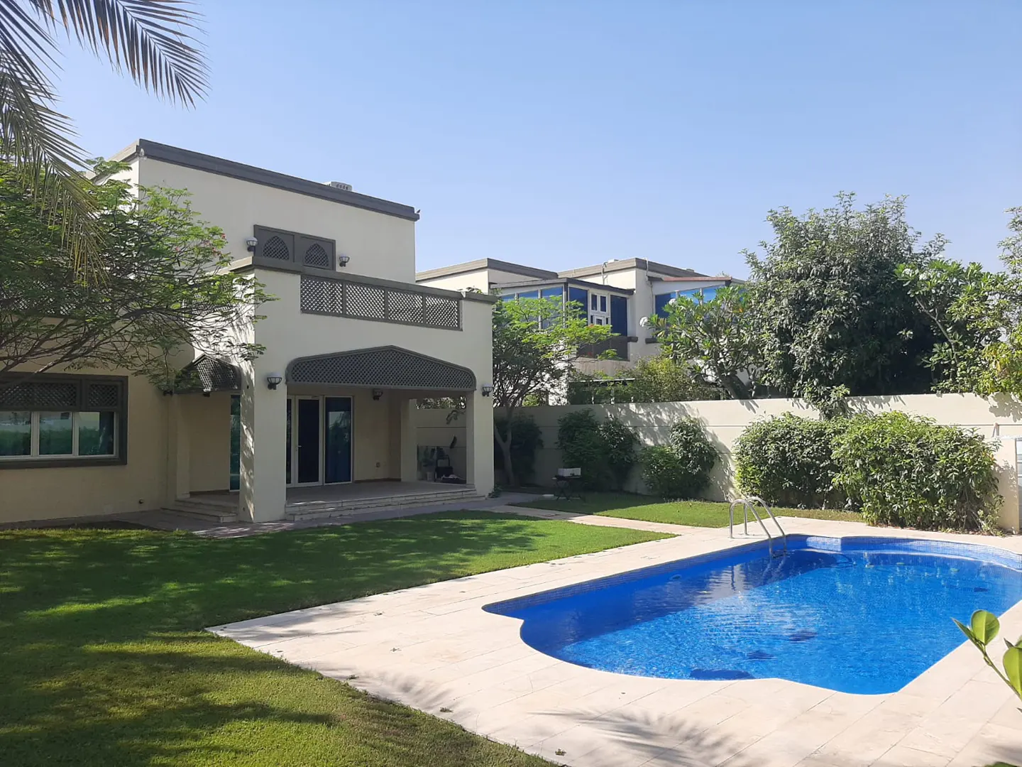 Beige two-story house with a balcony, green lawn, and a blue swimming pool on a sunny day.