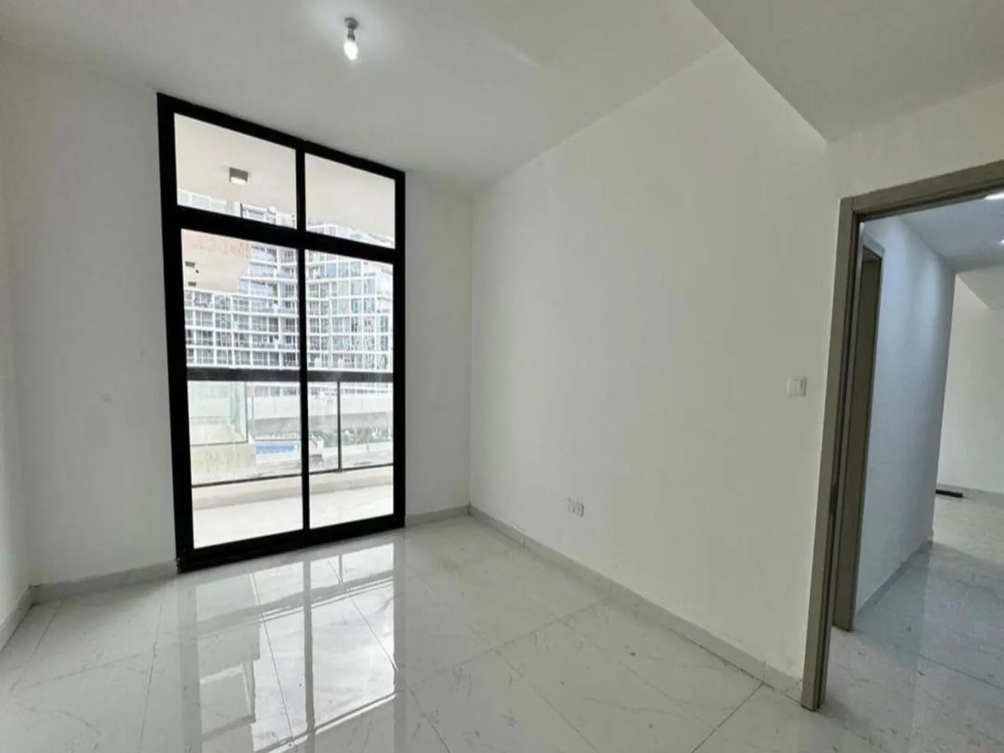 Bright, empty room with white walls and marble floors. A black-framed window shows a city view. An open doorway leads to another room.