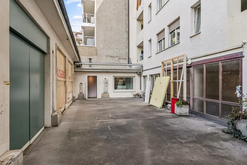 Exterior view of a building's courtyard with a concrete floor, white walls, and a green door on the left. Construction materials are stacked against the building on the right.