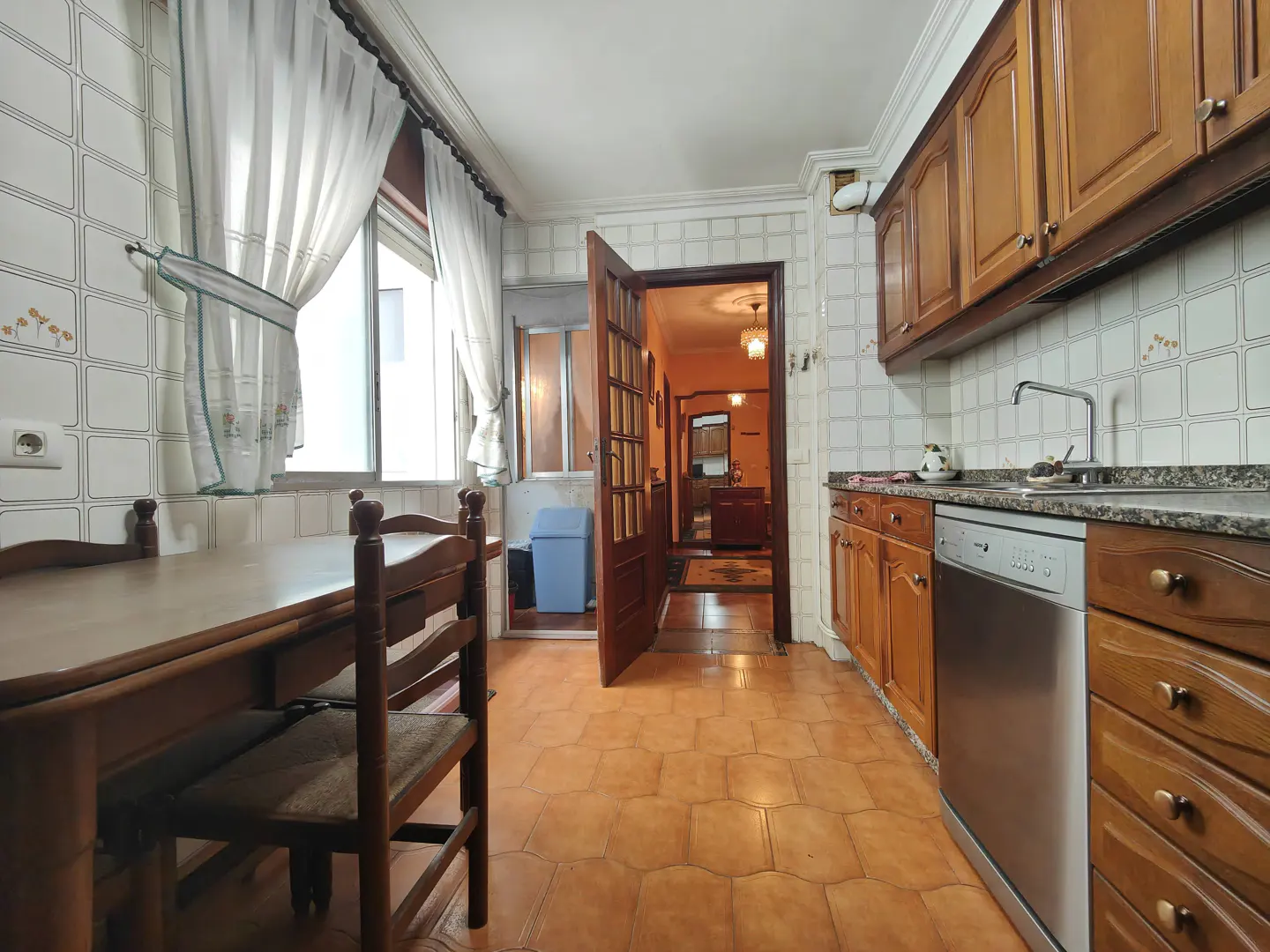 A kitchen with brown cabinets, a stainless steel dishwasher, and a wooden table with chairs. An open door leads to another room.