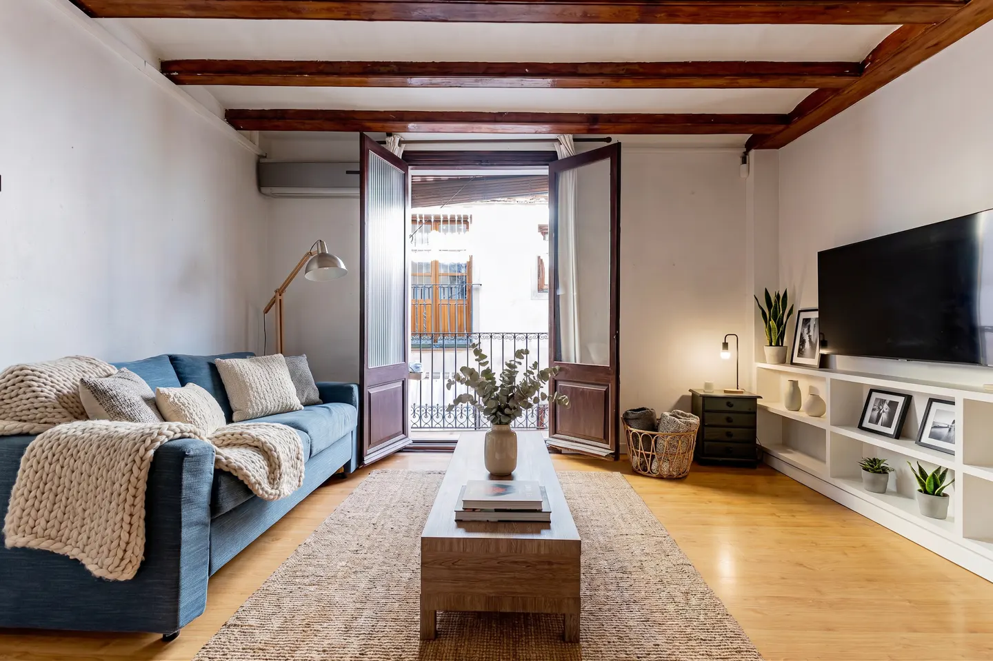Bright living room with blue sofa, wooden beams, and open doors to a balcony. A coffee table sits on a woven rug.