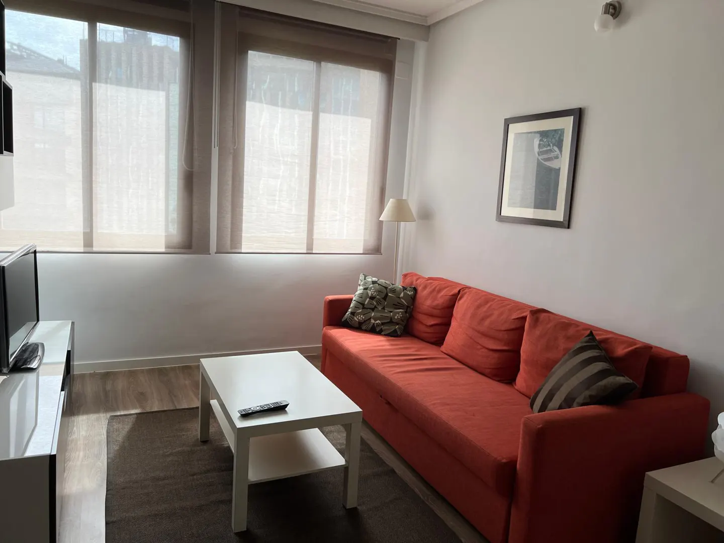 Living room with an orange sofa, white coffee table, and TV stand. Windows with blinds provide natural light. A framed picture hangs on the gray wall.