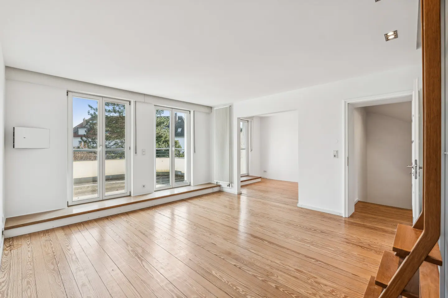 Bright, empty room with light wood floors, white walls, and large windows overlooking a balcony. A doorway leads to another room.