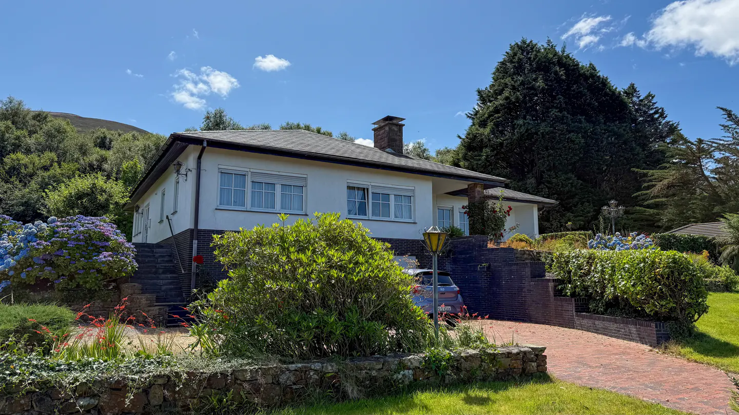 A white, single-story house with a gray roof, chimney, and manicured landscaping on a sunny day.