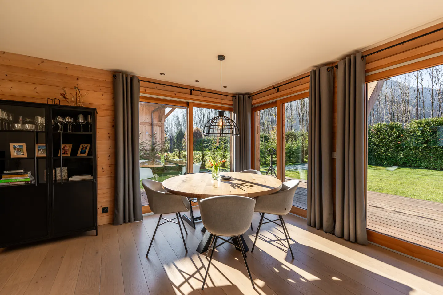 Bright dining room with wood walls, light wood floor, round table with gray chairs, and large windows overlooking a green lawn.