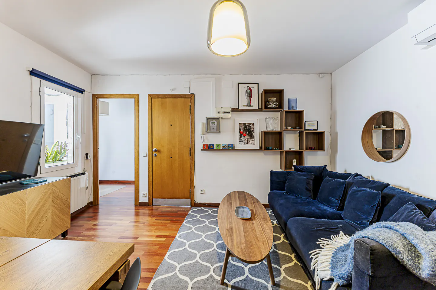 Bright living room with wood floors, a blue velvet sofa, and a patterned gray rug. A wooden coffee table sits in front of the sofa. Shelves and artwork decorate the white walls.